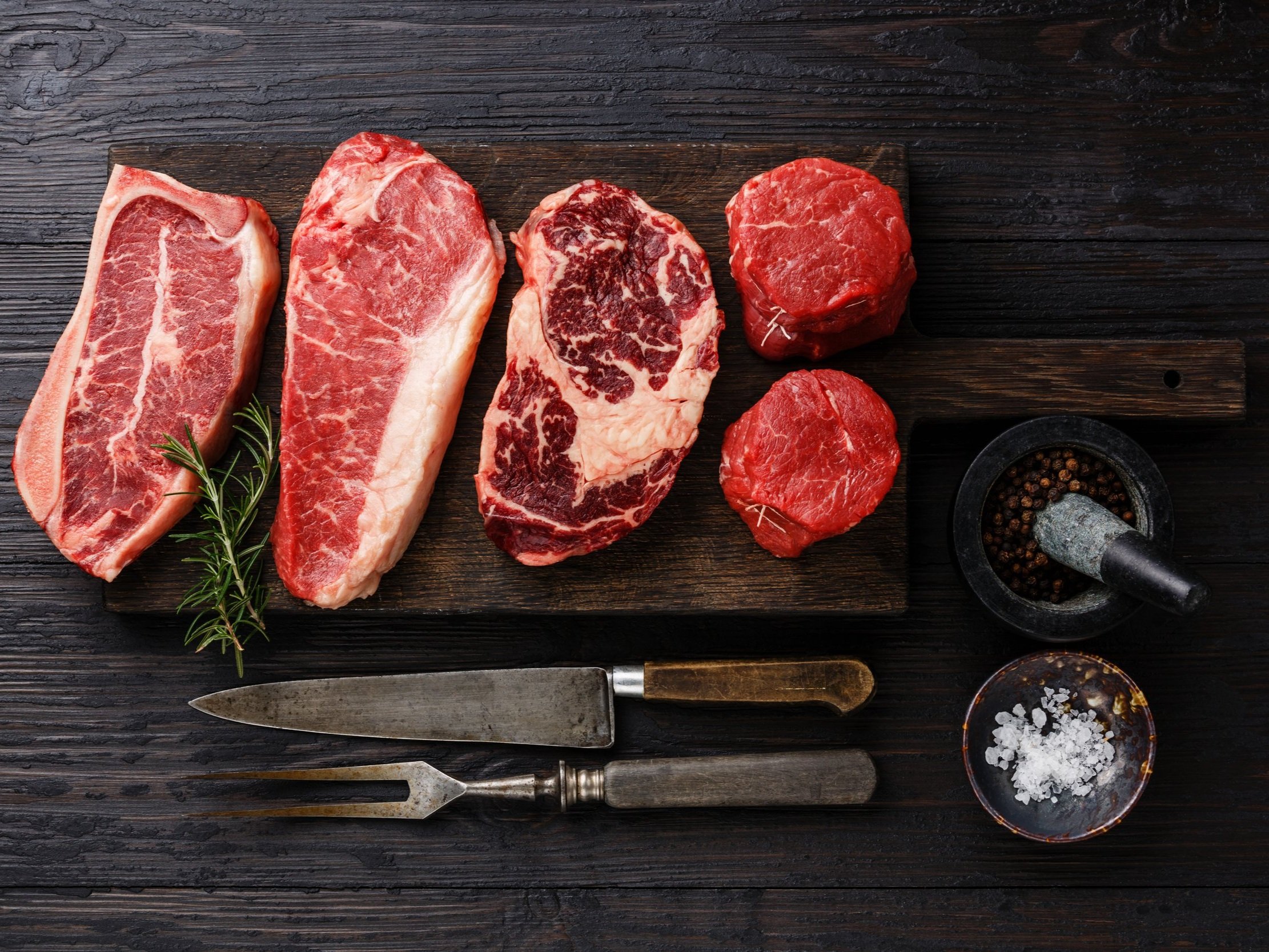 Assorted cuts of raw beef including ribeye, sirloin, and filet mignon displayed on a wooden cutting board, accompanied by fresh rosemary, a mortar and pestle with peppercorns, and a bowl of sea salt, perfect for grilling or cooking.