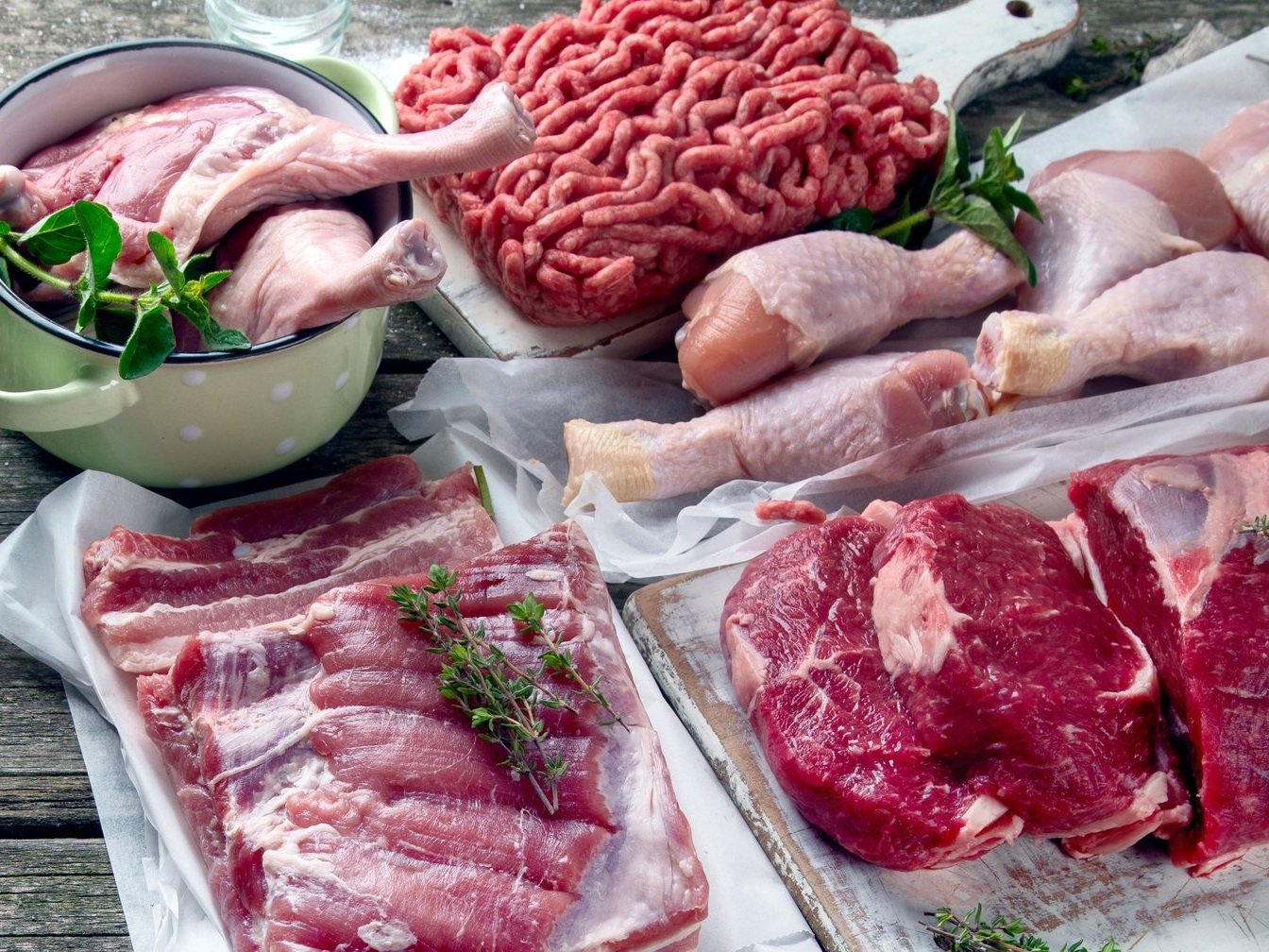 Assorted raw meats including chicken legs, ground beef, pork belly, and beef cuts, garnished with fresh herbs on a wooden surface.
