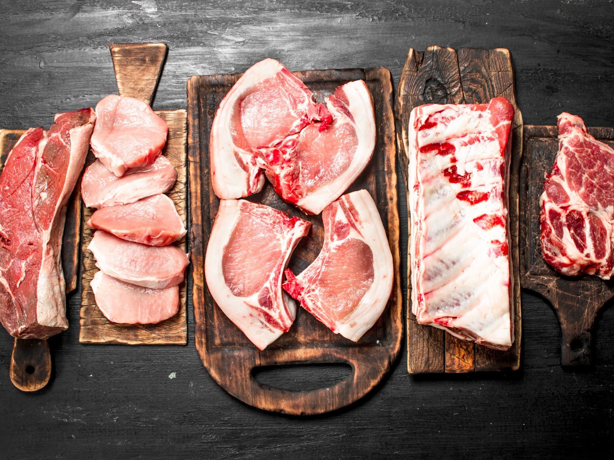 Assorted cuts of raw pork meat displayed on wooden cutting boards, including pork chops, ribs, and tenderloin, against a dark background. Ideal for showcasing fresh meat options for culinary use or recipes.